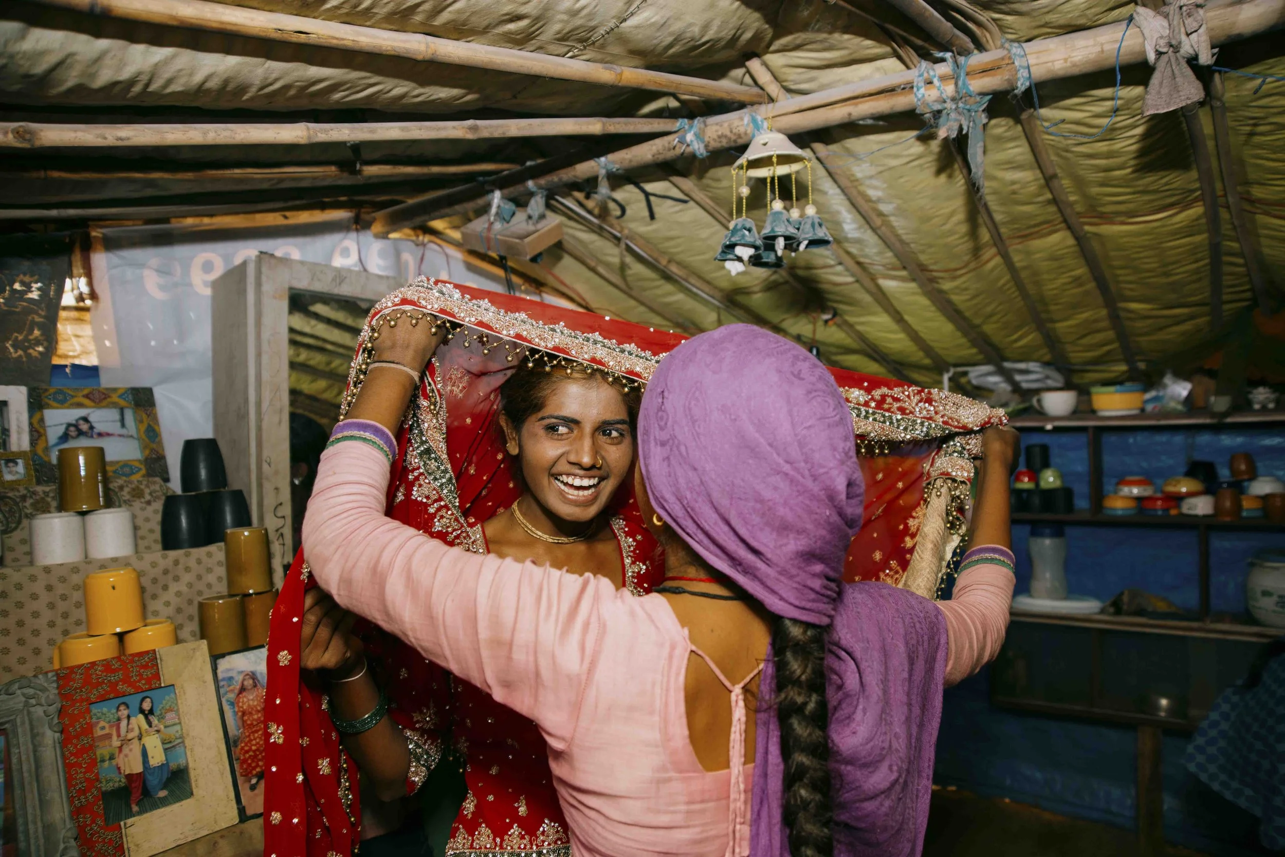 A woman in a traditional red bridal outfit with intricate embroidery is being helped into her wedding saree by another woman wearing purple, inside a modest home or shop with a bamboo and cloth roof, and shelves with various objects and photographs i