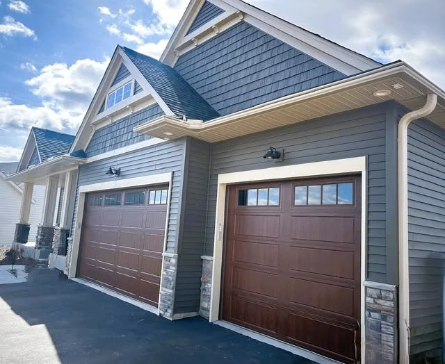 Photo of attached garages on a modern house with dark brown doors, gray siding, and stone accents, set against a partly cloudy sky.