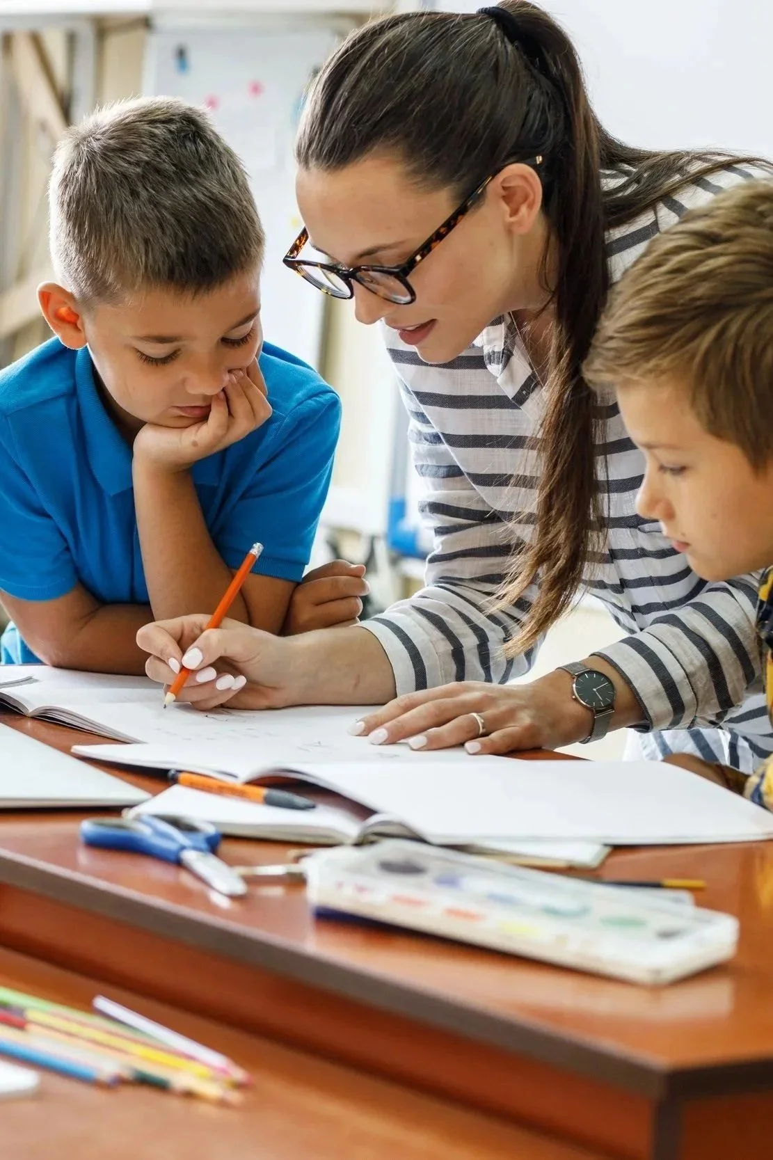 A teacher explaining something to two young boys at a classroom desk, with open notebooks, writing utensils, and art supplies on the desk.