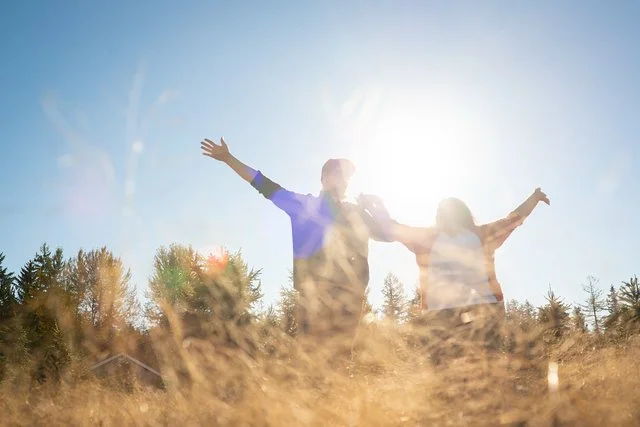 Two people with arms raised standing outdoors in a field, with the sun shining behind them and a clear blue sky.