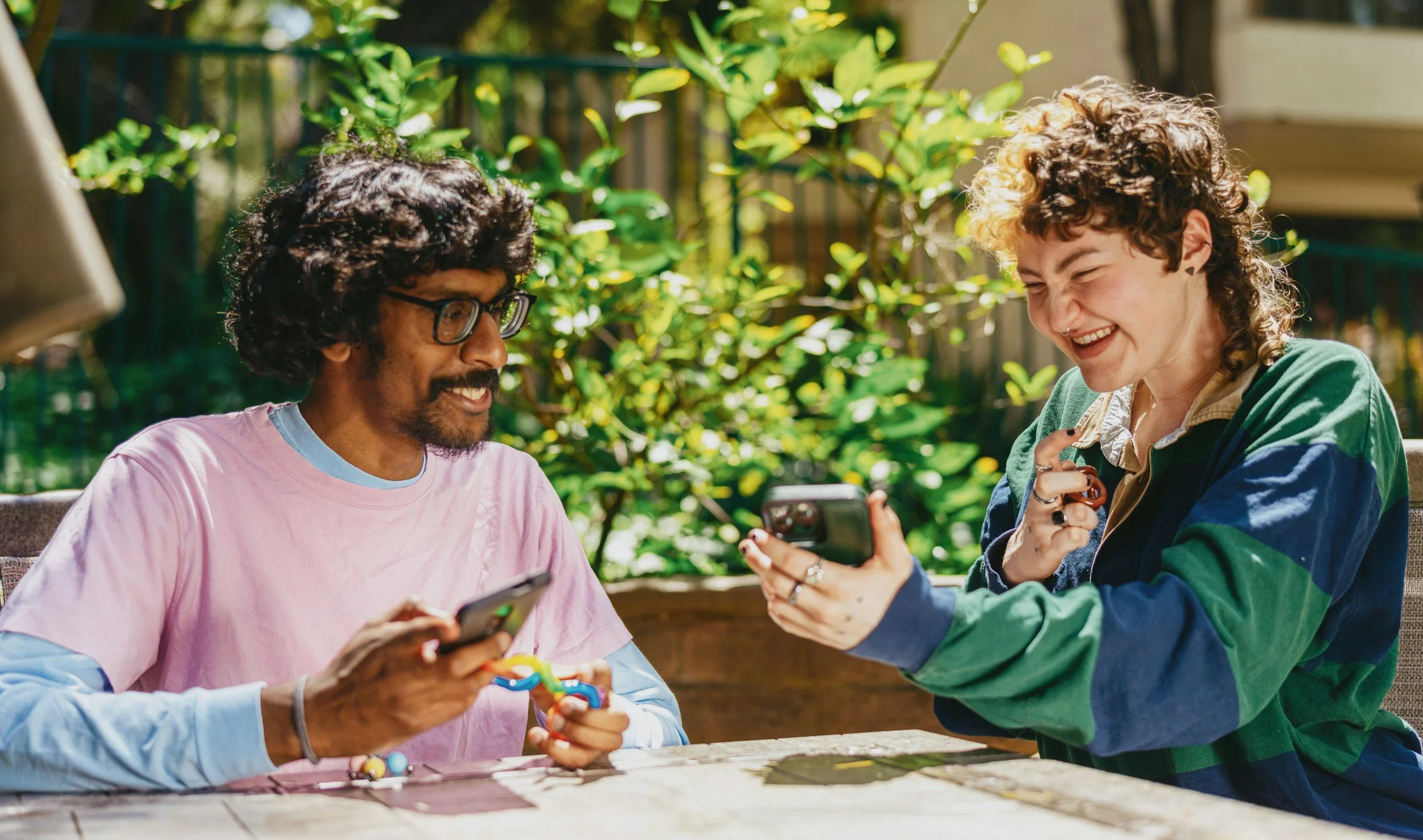 Two friends sitting at a table outdoors, laughing and looking at their phones, surrounded by green plants.