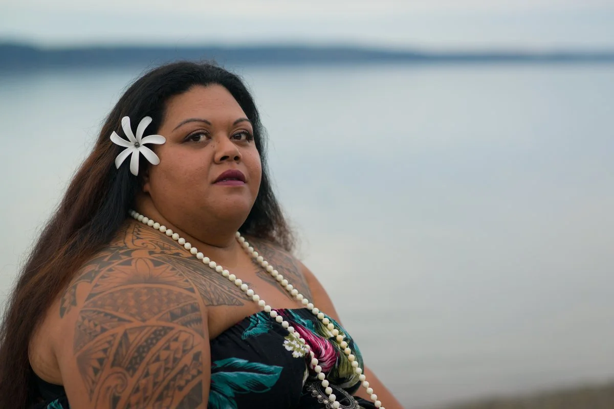 A woman with long dark hair, a large white flower in her hair, and tattoos on her arm, wearing a floral strapless top and a pearl necklace, standing by a body of water.