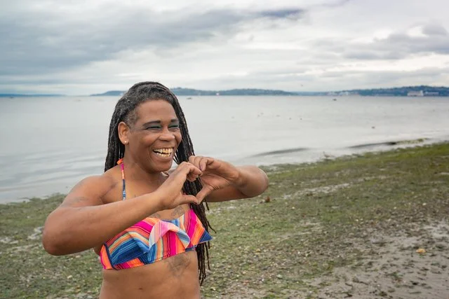 A woman smiling and making a heart shape with her hands on a beach with water and cloudy sky in the background.