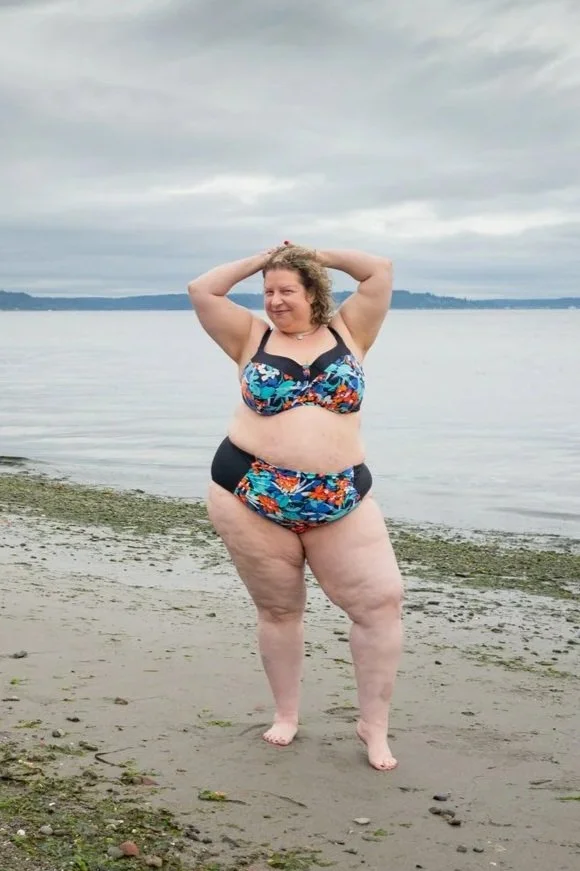 A woman in a colorful bikini standing on a sandy beach with her hands behind her head, smiling, with water and a cloudy sky in the background.