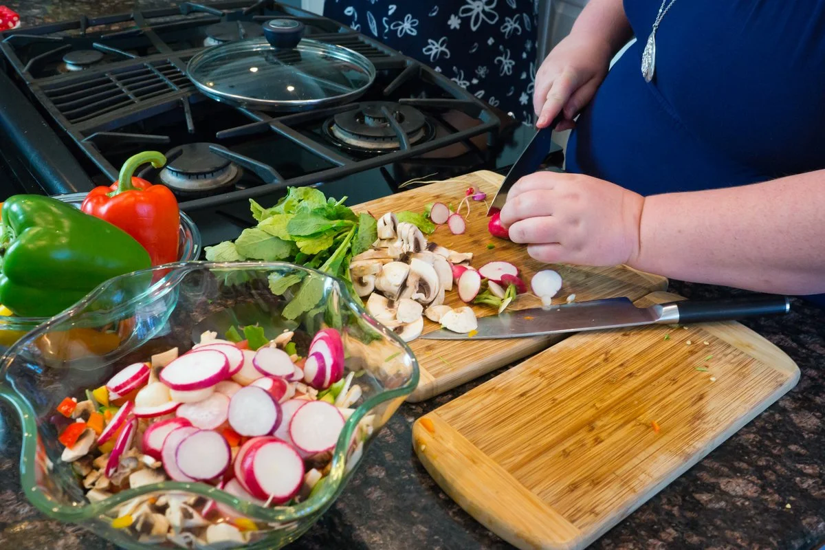 Person chopping radishes, mushrooms, and greens on a wooden cutting board in a kitchen, with colorful bell peppers and a glass dish of sliced radishes nearby.