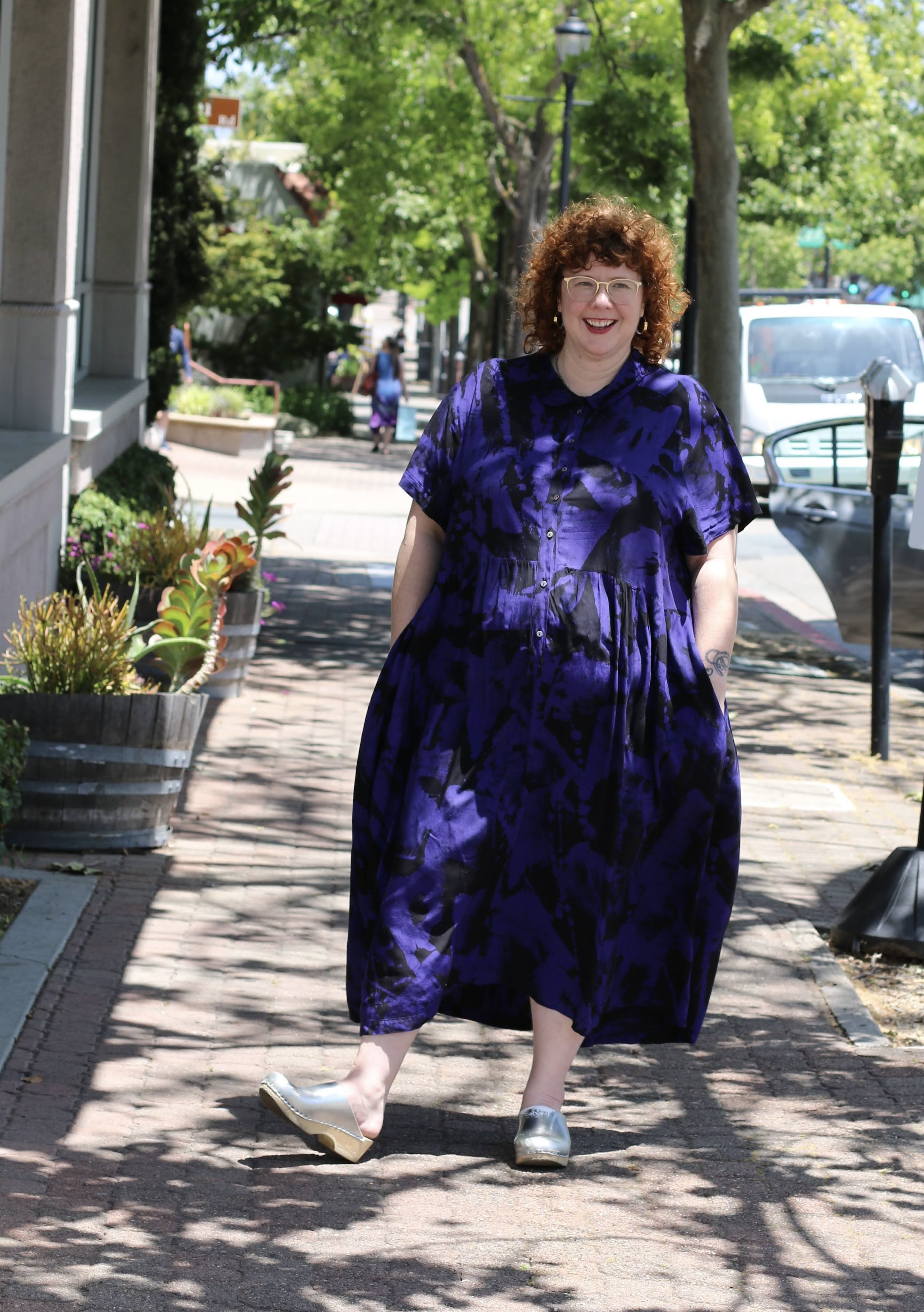 A woman with curly red hair, glasses, and a broad smile, walking on a sunny sidewalk lined with potted plants and trees. She is wearing a purple and black patterned dress and metallic silver shoes.