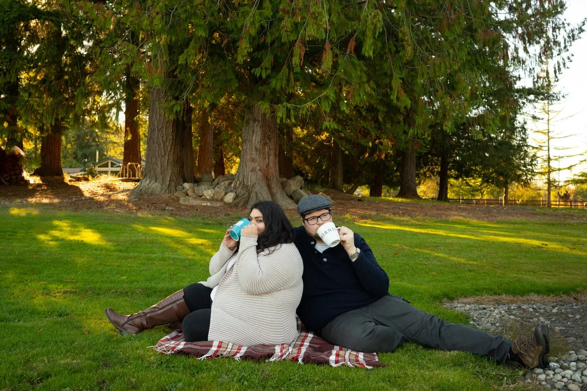 A man and a woman sitting on a picnic blanket on grass, drinking from mugs, with large trees and a sunset in the background.