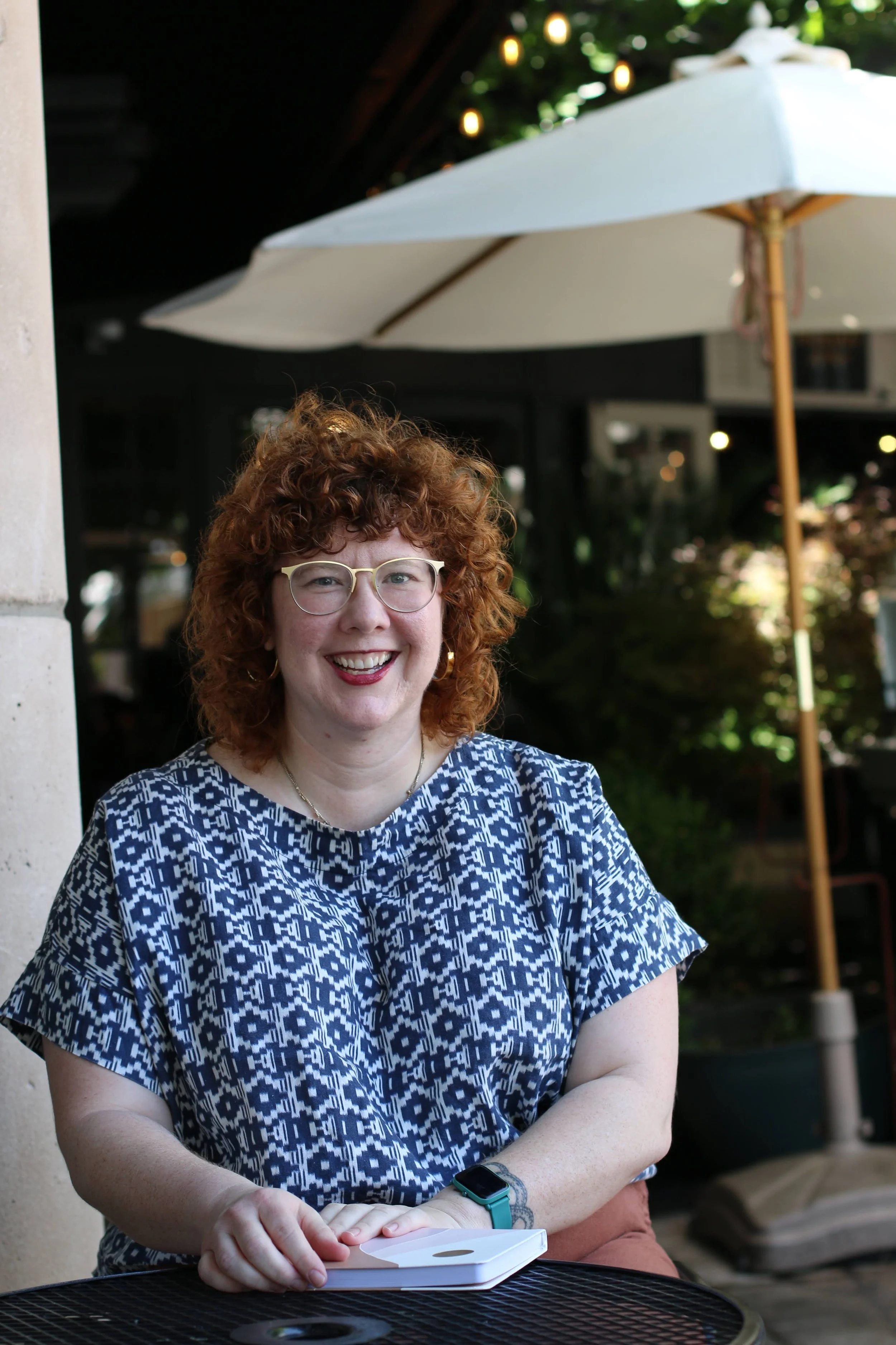 Smiling woman with curly red hair and glasses sitting at an outdoor table with a tablet, under a large white umbrella.