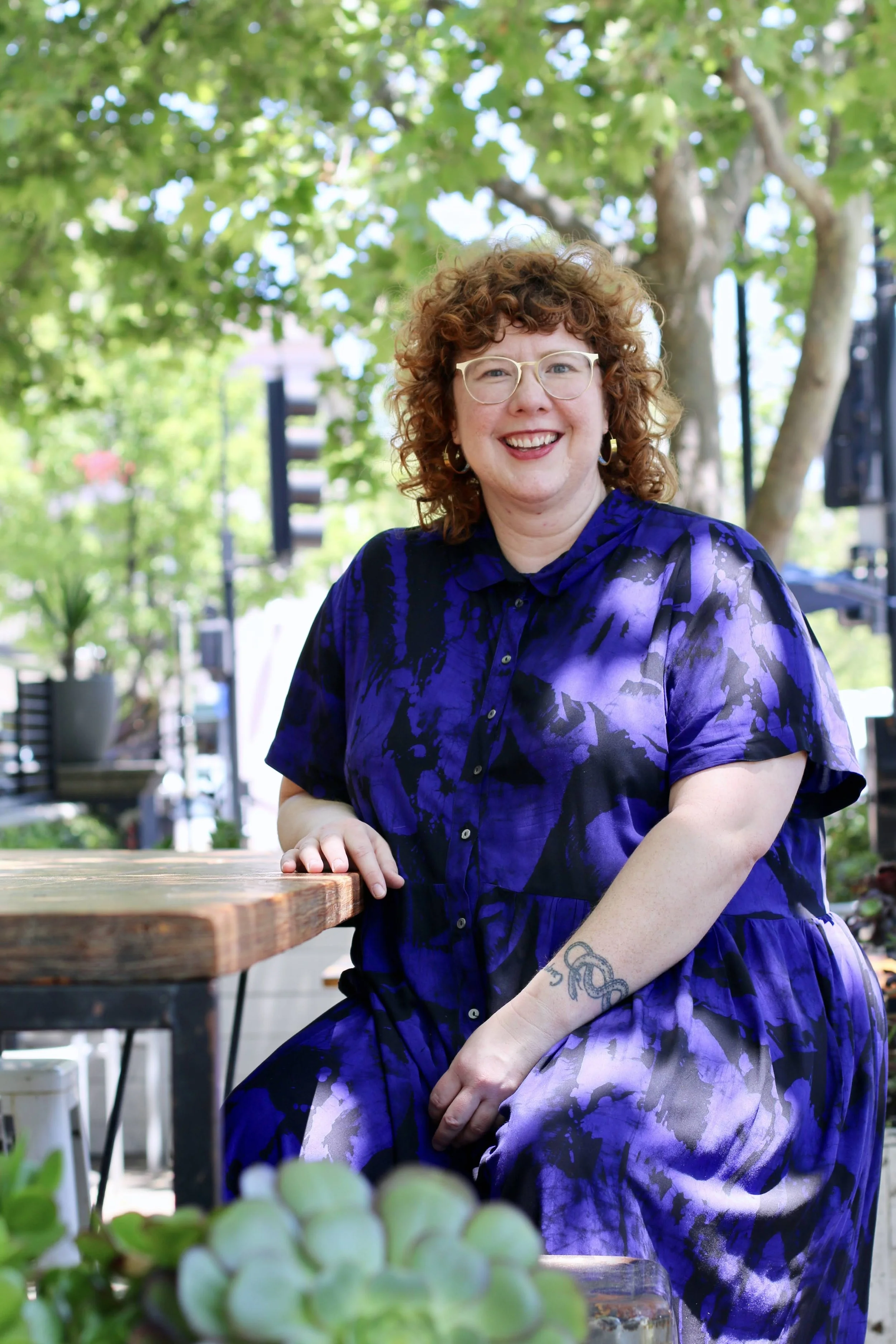 A woman with curly red hair sitting outdoors under a leafy green tree, smiling, wearing glasses and a black and purple patterned dress.