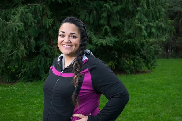 A woman with dark hair styled in a braid, smiling outdoors, wearing a black and purple athletic jacket, with green trees and grass in the background.