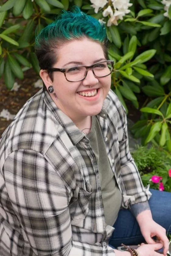 Woman with teal and brown hair, glasses, and a nose piercing smiling outdoors, surrounded by greenery and white flowers.