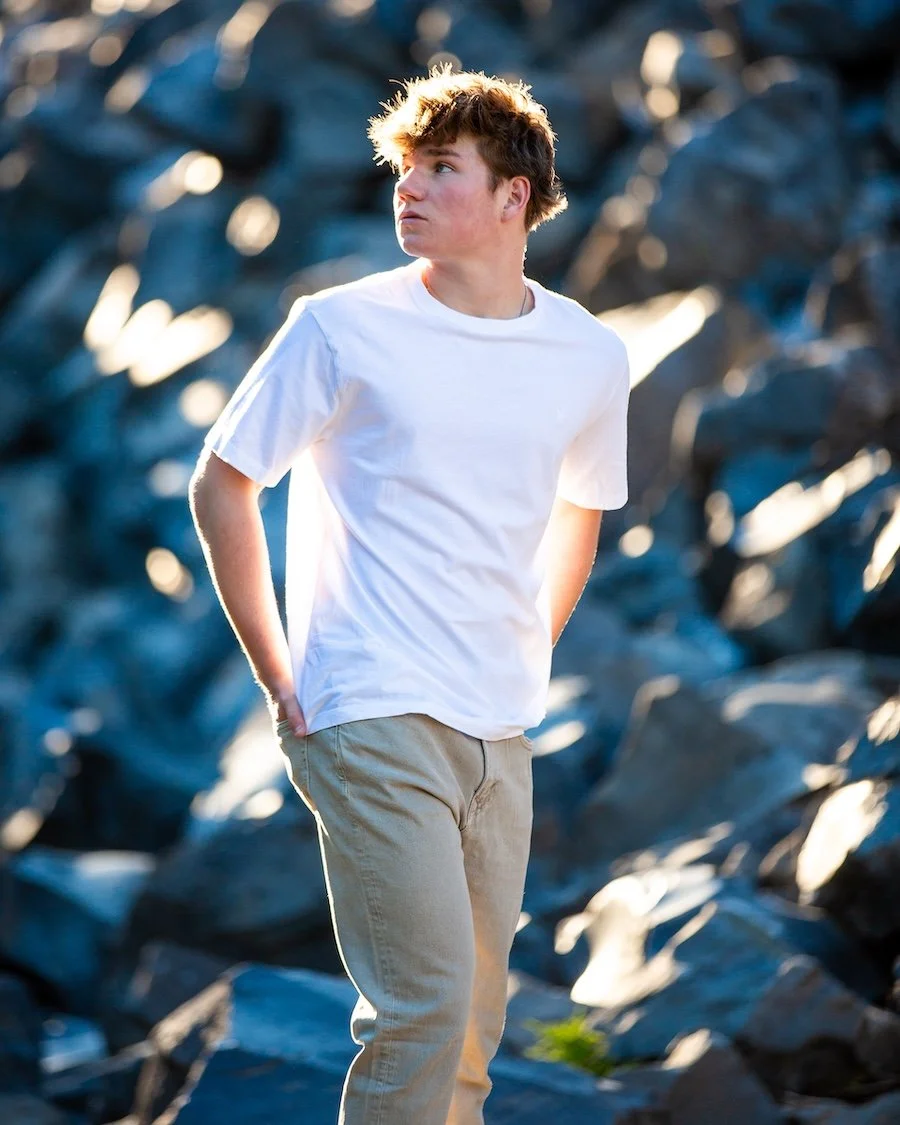 A young man with curly brown hair wearing a white t-shirt and beige pants, standing outdoors with rocks in the background and sunlight illuminating his head from behind.