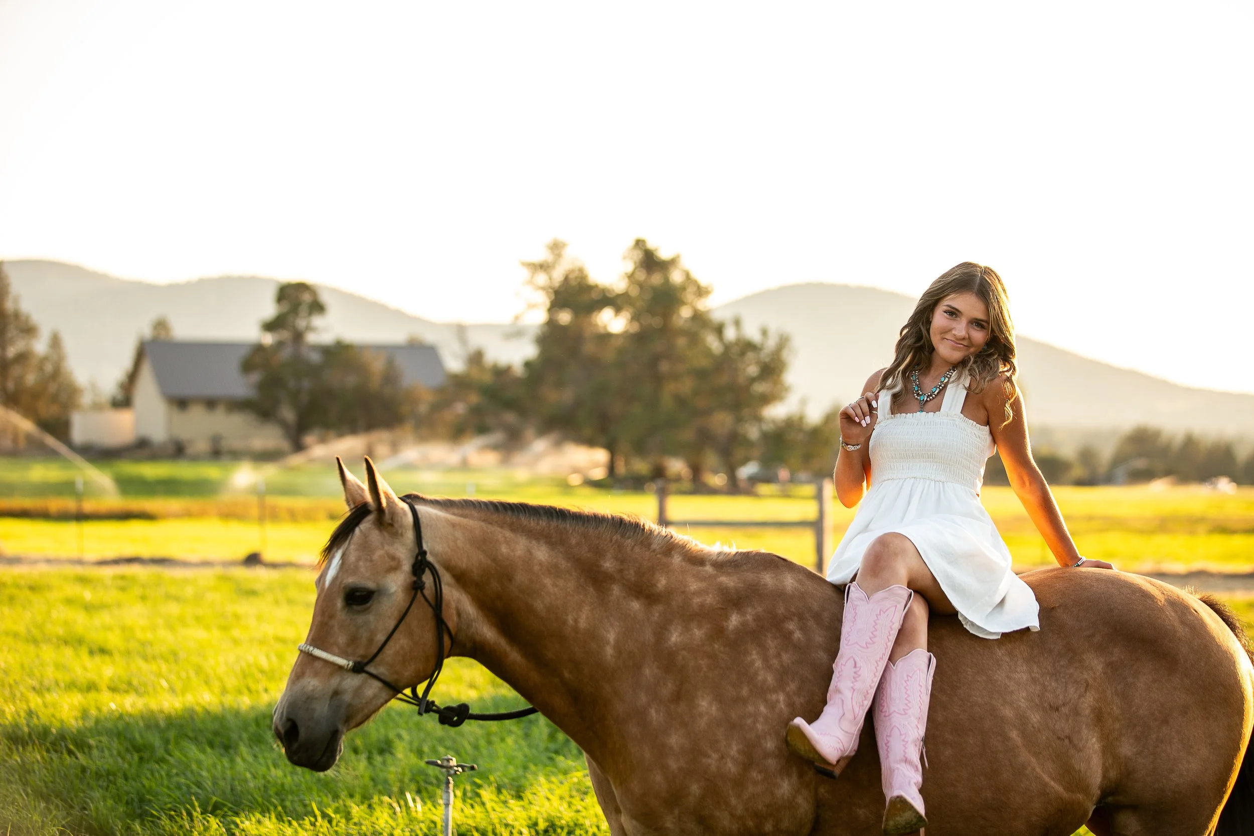 A young woman in a white dress and pink cowboy boots sitting on a tan horse in a grassy field at sunset.