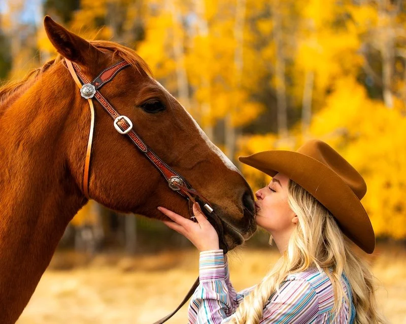 A woman with long blonde hair, wearing a brown cowboy hat and a colorful striped shirt, is kissing a brown horse on its nose in a fall landscape with yellow trees.