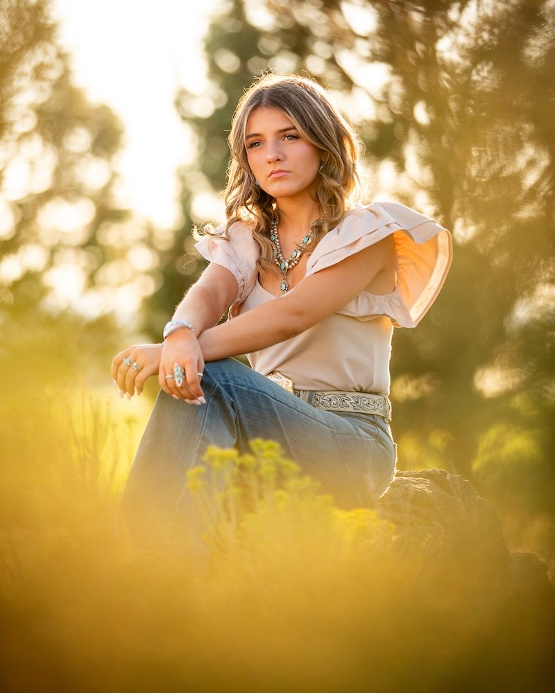 Young woman with wavy blonde hair sitting outdoors in a field during sunset, wearing a cream-colored blouse with ruffled sleeves and multiple necklaces, looking contemplative.