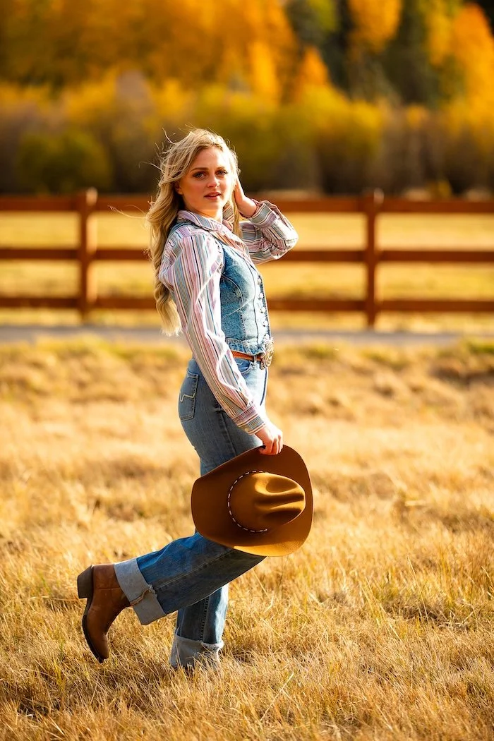 A young woman in a plaid shirt, denim vest, jeans, and cowboy boots holding a brown cowboy hat, standing in a field with autumn trees and a wooden fence in the background.
