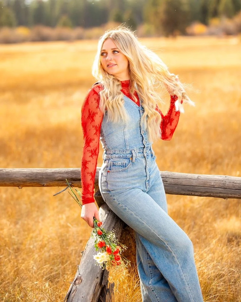 A young woman with long blonde hair wearing a red lace long-sleeve shirt and denim overalls, standing outdoors in a golden field during sunset, holding a small bouquet of flowers.