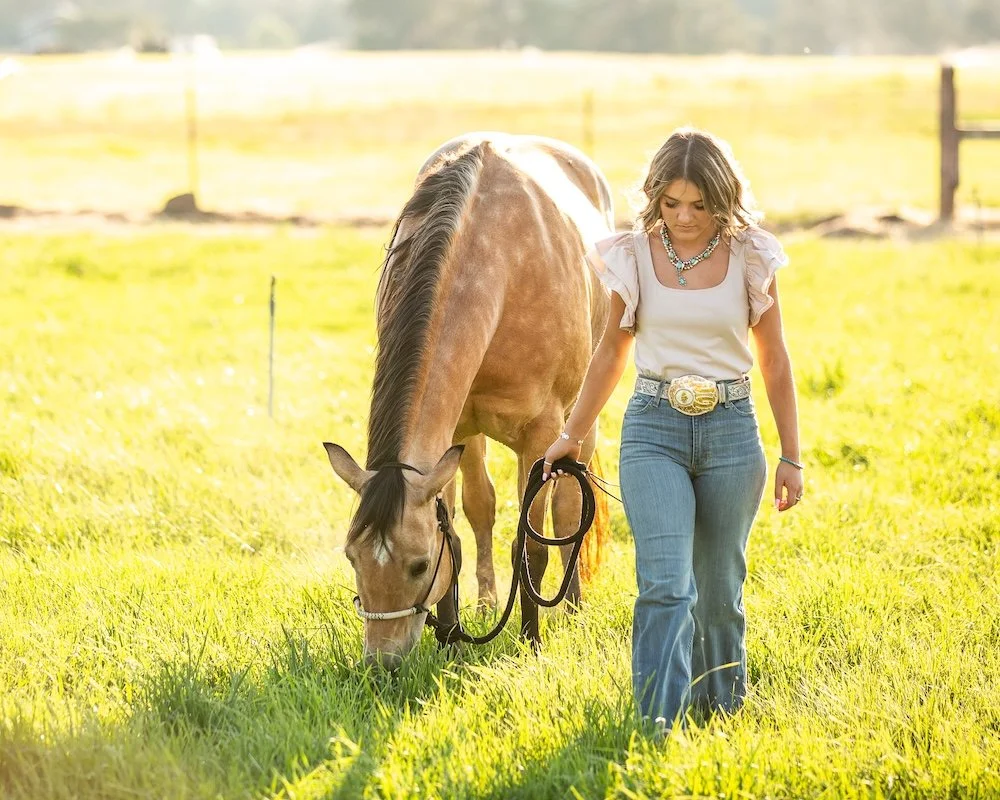 A woman walking a brown horse in a sunlit grassy field, holding the horse's reins.