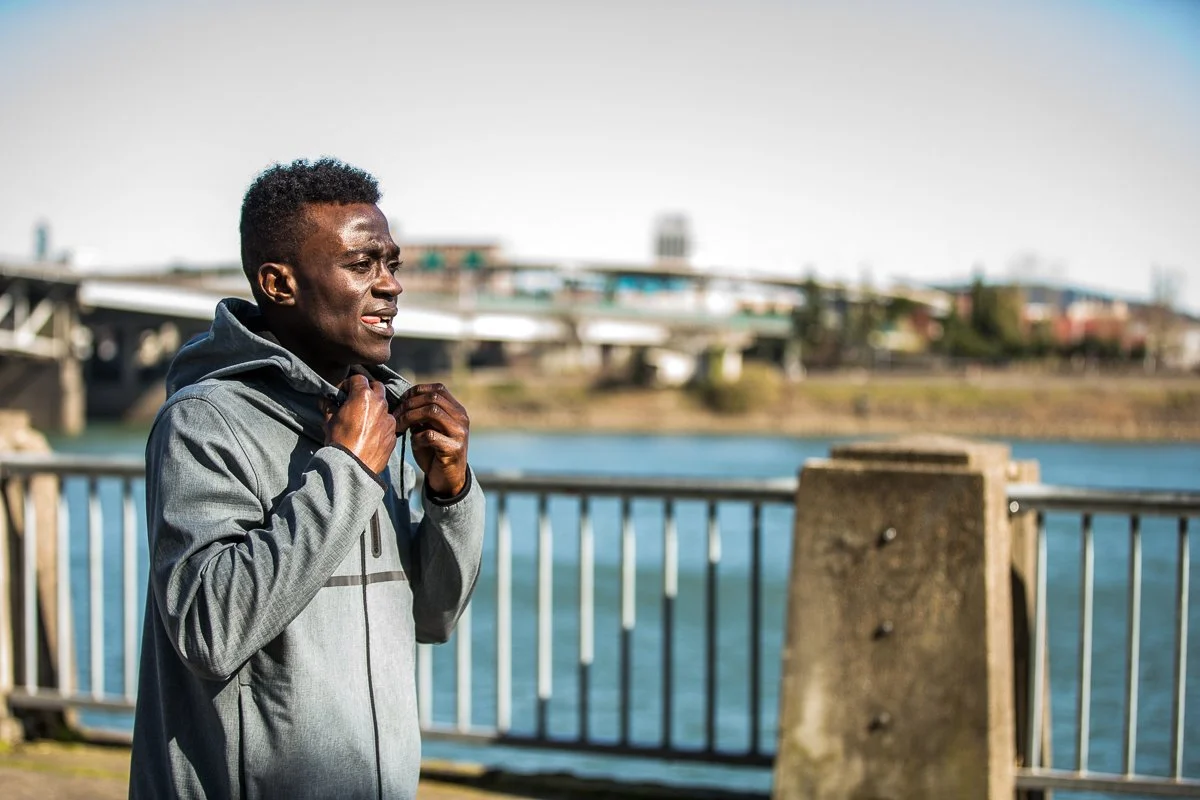 Man in athletic jacket jogging outdoors near water with bridge and cityscape in background