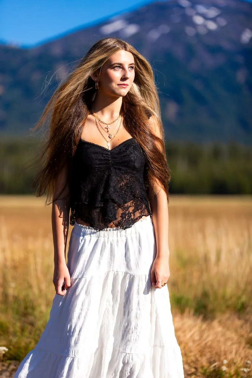 A young woman with long, light brown hair stands outdoors in a field with a mountain and a clear blue sky in the background. She is wearing a black lace sleeveless top and a long white skirt, accessorized with layered necklaces and hoop earrings.
