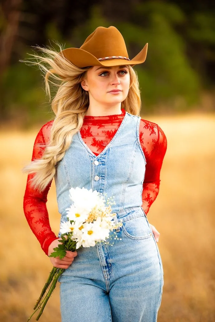 A young woman with long blonde hair, wearing a cowboy hat, a red lace long-sleeve top, and denim overalls, holding a bouquet of daisies in a field with trees in the background.
