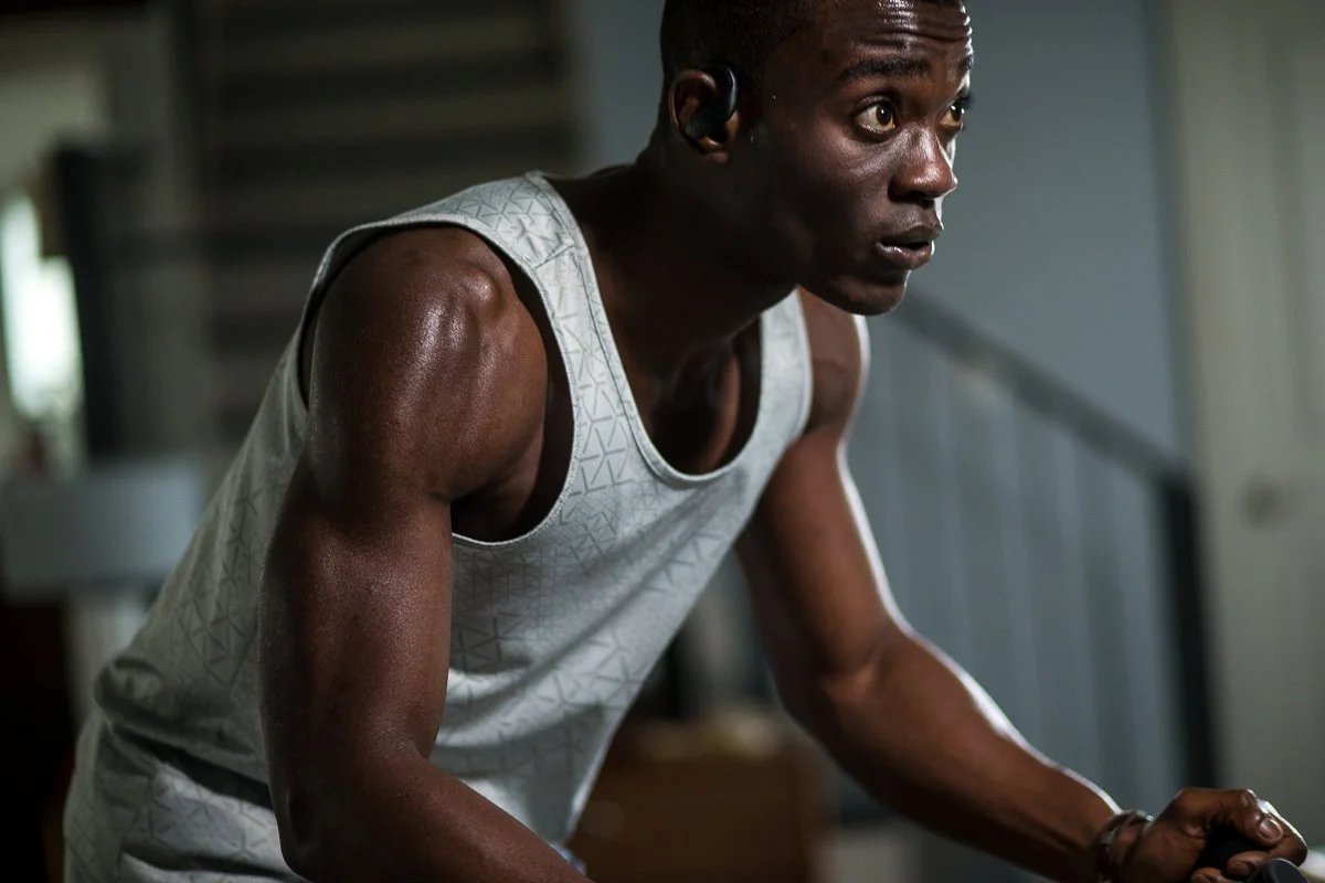 A man in athletic attire and a wireless earbud, leaning forward with a focused expression during a workout indoors.