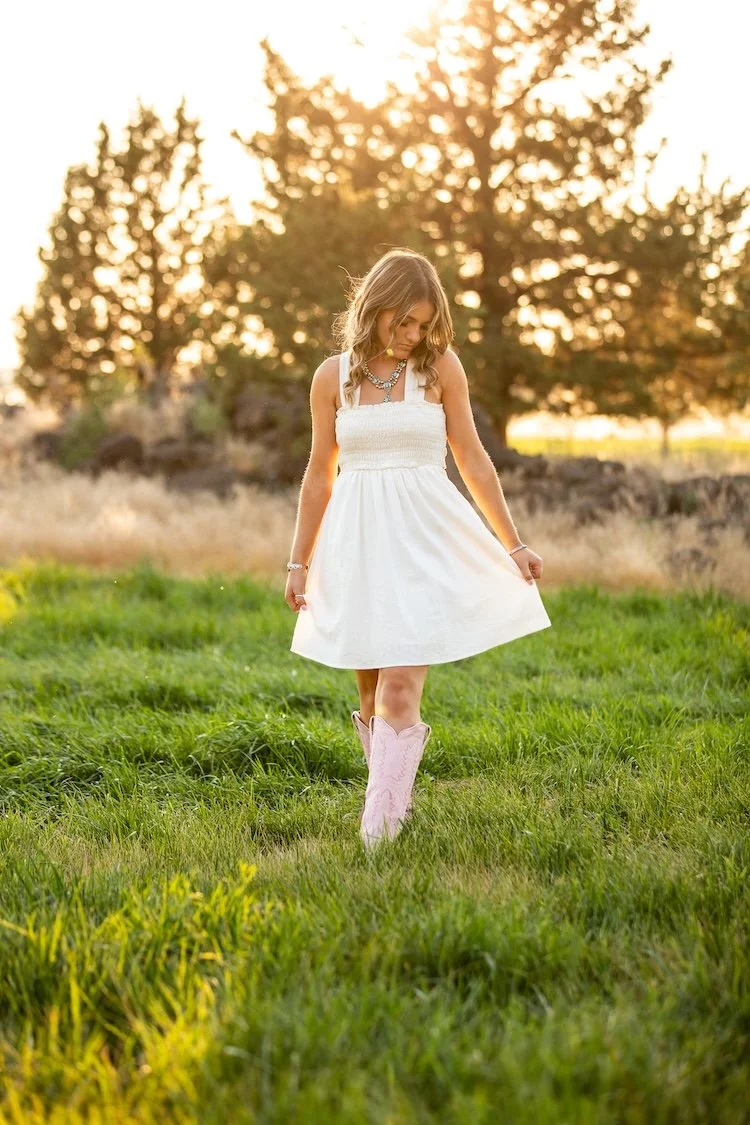 A young girl in a white dress and pink cowboy boots walking through a grassy field during sunset.