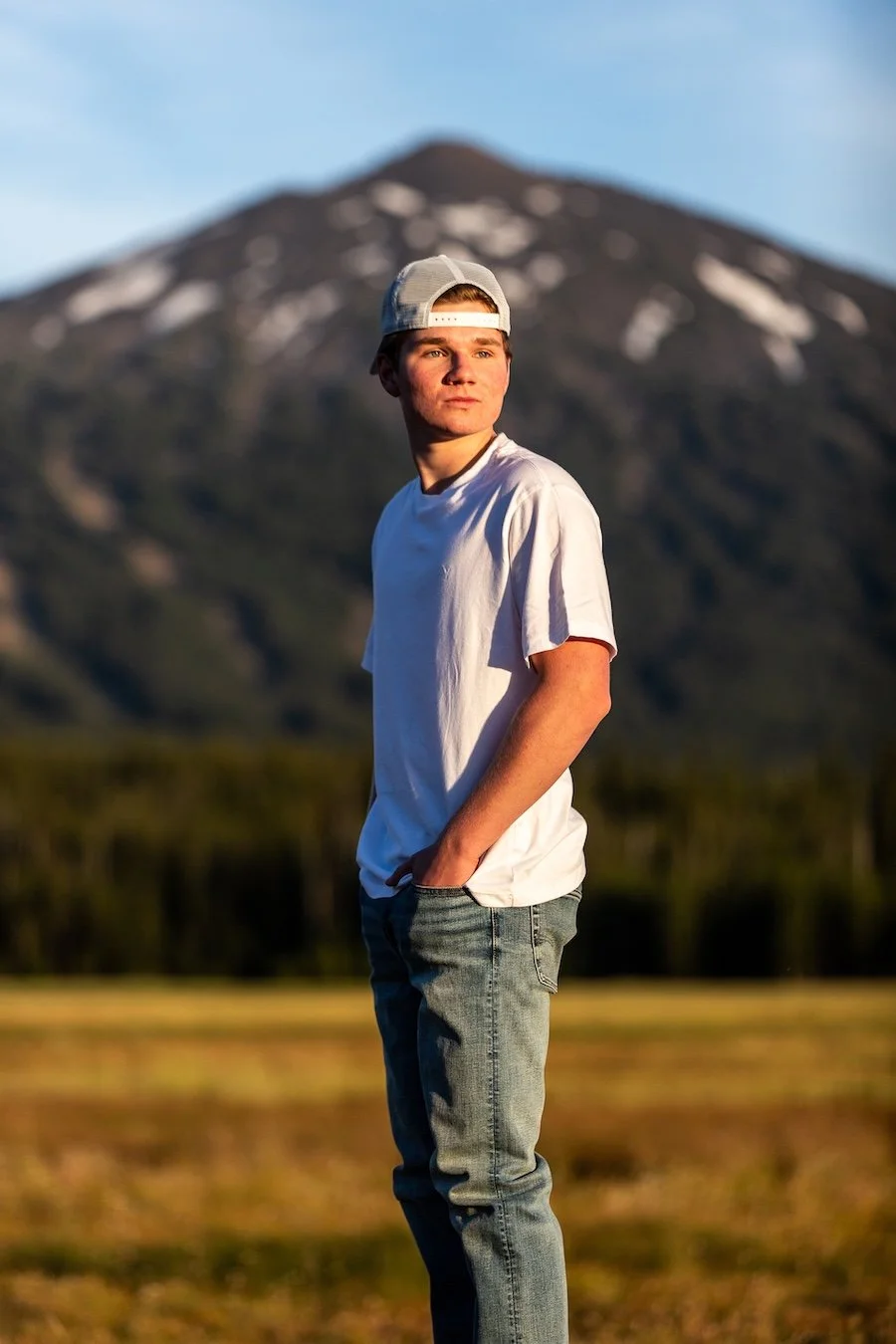 Young man in a white t-shirt and jeans standing outdoors with mountains in the background during sunset.