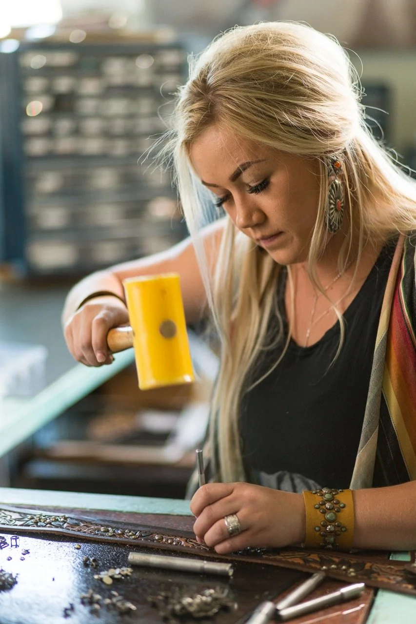 A woman working on a jewelry piece in a workshop, holding a yellow rubber mallet with tools and jewelry materials on the table.