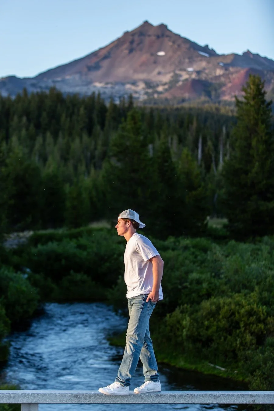 A young man in casual clothing, including a white t-shirt, jeans, and a backward baseball cap, walking along a narrow bridge over a river in a lush green forest with mountains in the background.