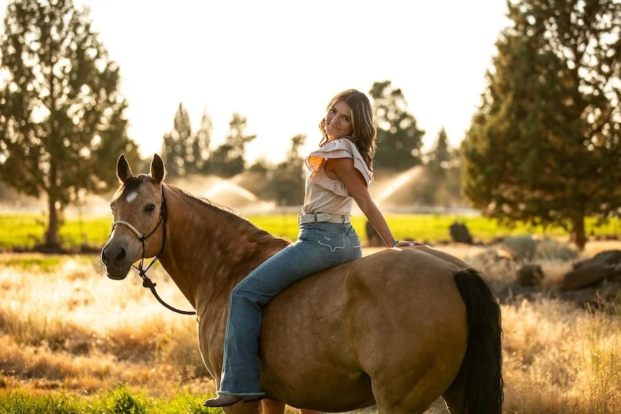 A woman sitting on a brown horse in a rural outdoor setting during sunset, with trees and a yellow field in the background.