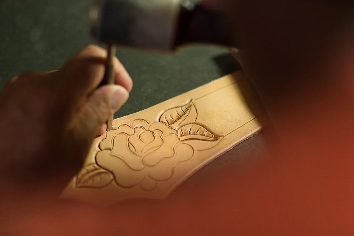 Close-up of a person's hand carving a floral design on a piece of leather with a carving tool.