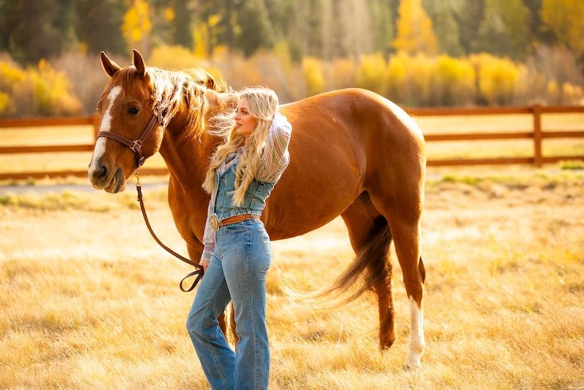 A young woman with long blonde hair standing beside a chestnut horse in a field during autumn. The woman is wearing jeans and a vest, and the horse has a leather halter. There is a wooden fence and colorful fall trees in the background.