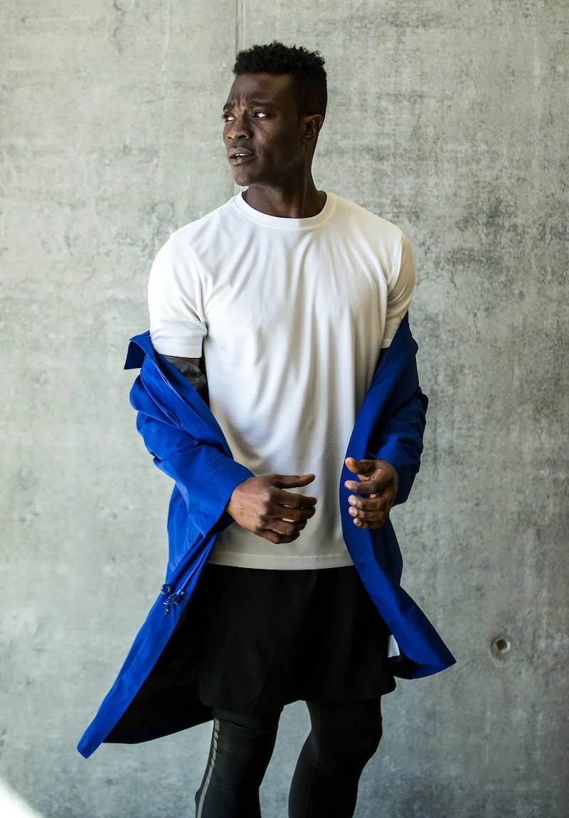A man with short curly hair wearing a white t-shirt, black athletic pants, and a blue jacket draped over his shoulders, standing against a gray concrete wall, looking to his left with a serious expression.
