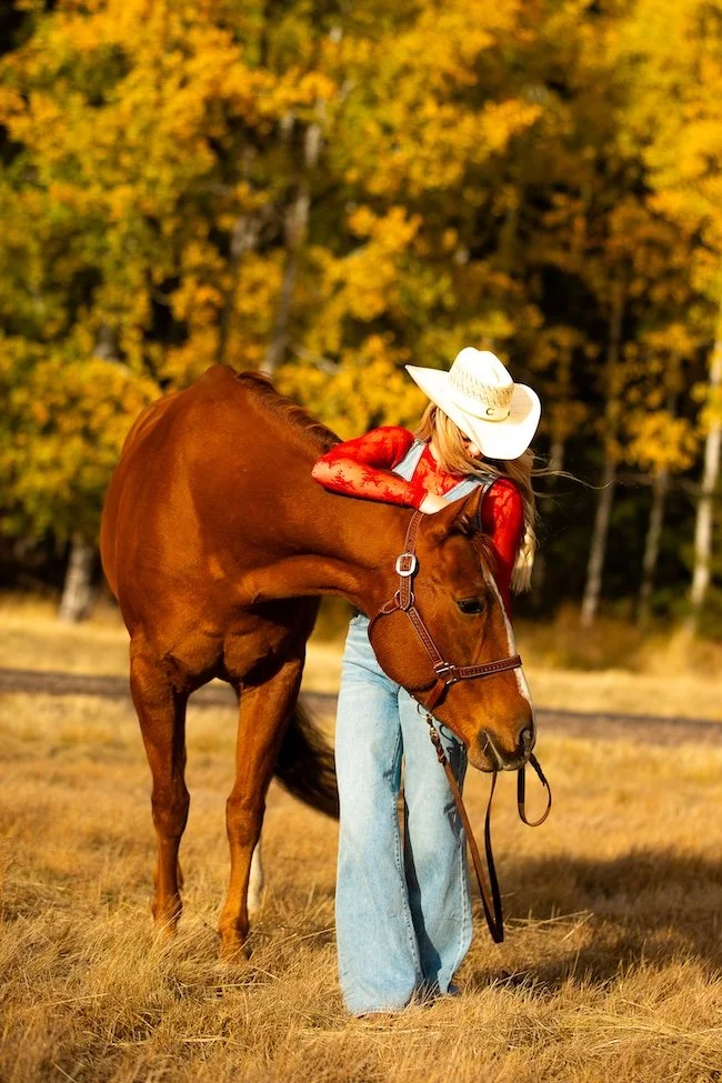 A woman wearing a white cowboy hat, red long-sleeve top, and light blue jeans hugging a brown horse in a field with autumn-colored trees in the background.