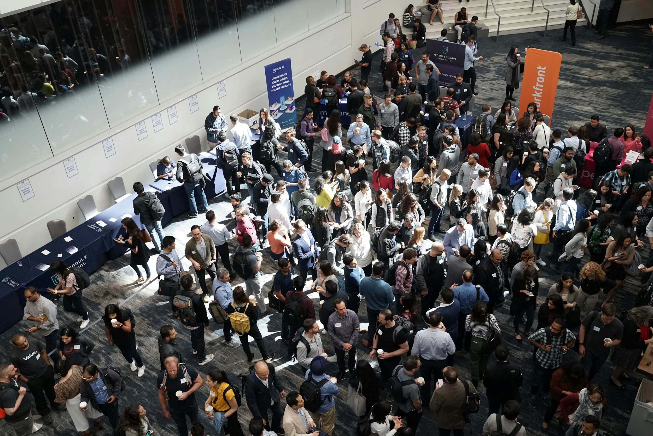 Crowd of people attending an event or conference, standing in line or walking through the venue, with registration desks, banners, and informational tables visible.