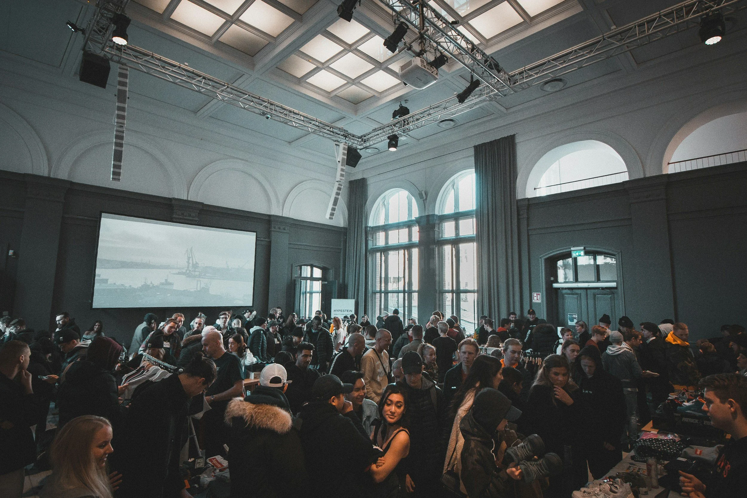 Crowd of people inside a large, upscale event space with tall arched windows and a large screen at the front.