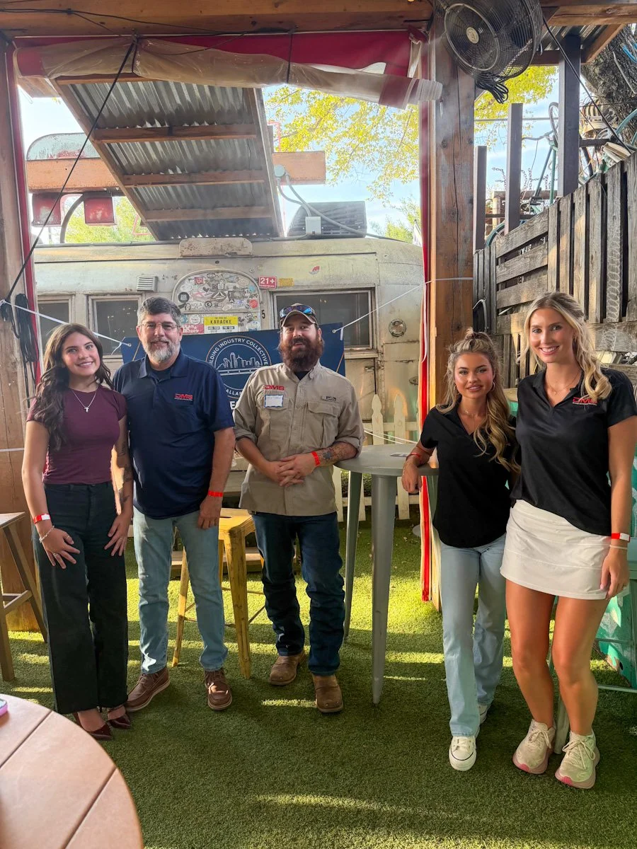 Group of five people standing in a semi-outdoor space with a vintage trailer in the background, posing for a photo, with trees and outdoor sunlight visible.