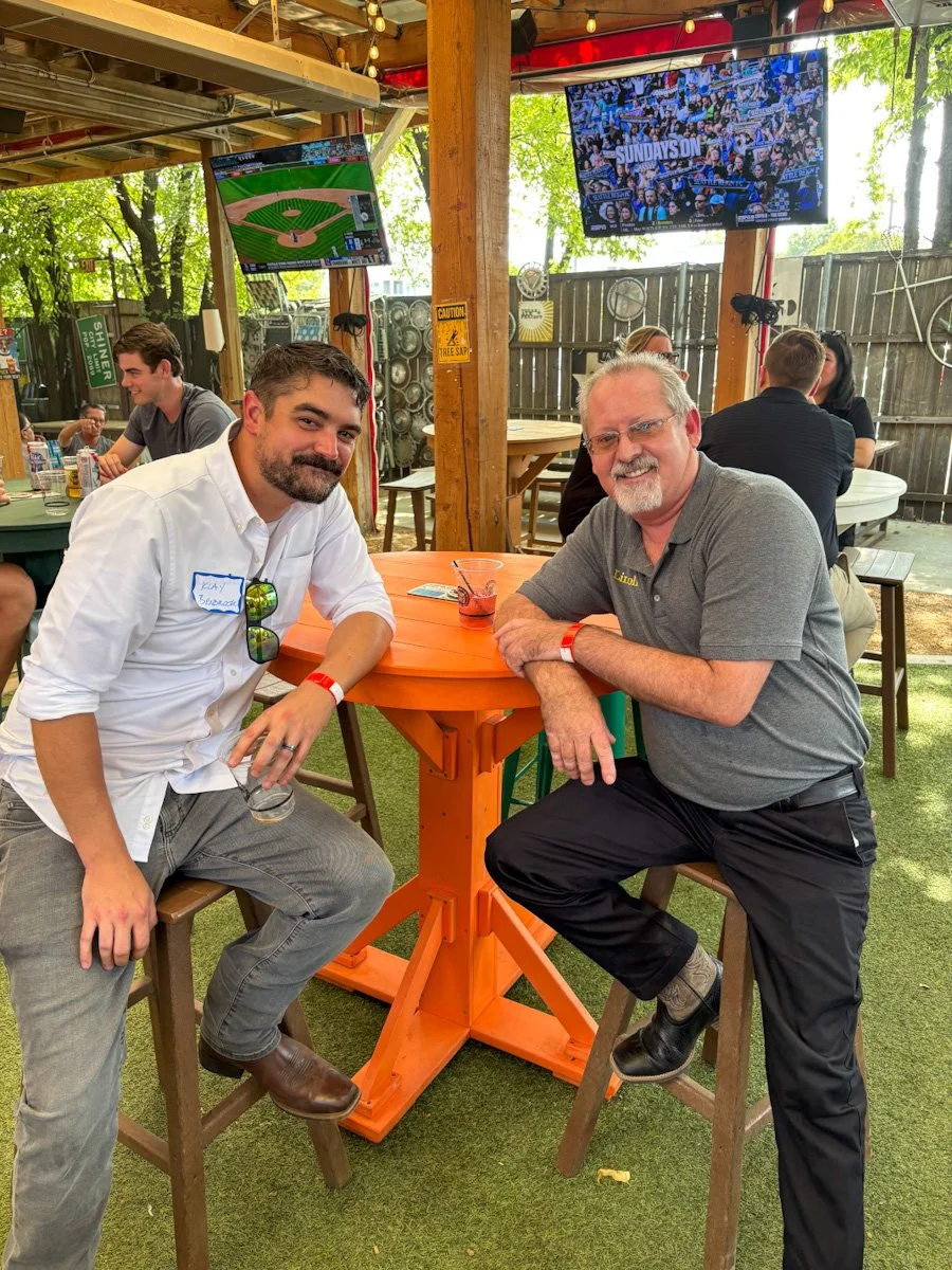 Two men sitting at a tall orange table outdoors at a bar or restaurant. One man has a beard, wearing a white shirt with sunglasses hanging on his chest. The other man has gray hair, glasses, and a gray polo shirt. Behind them are other patrons and two TVs showing baseball and a news program. The setting has green artificial grass, trees, and string lights.