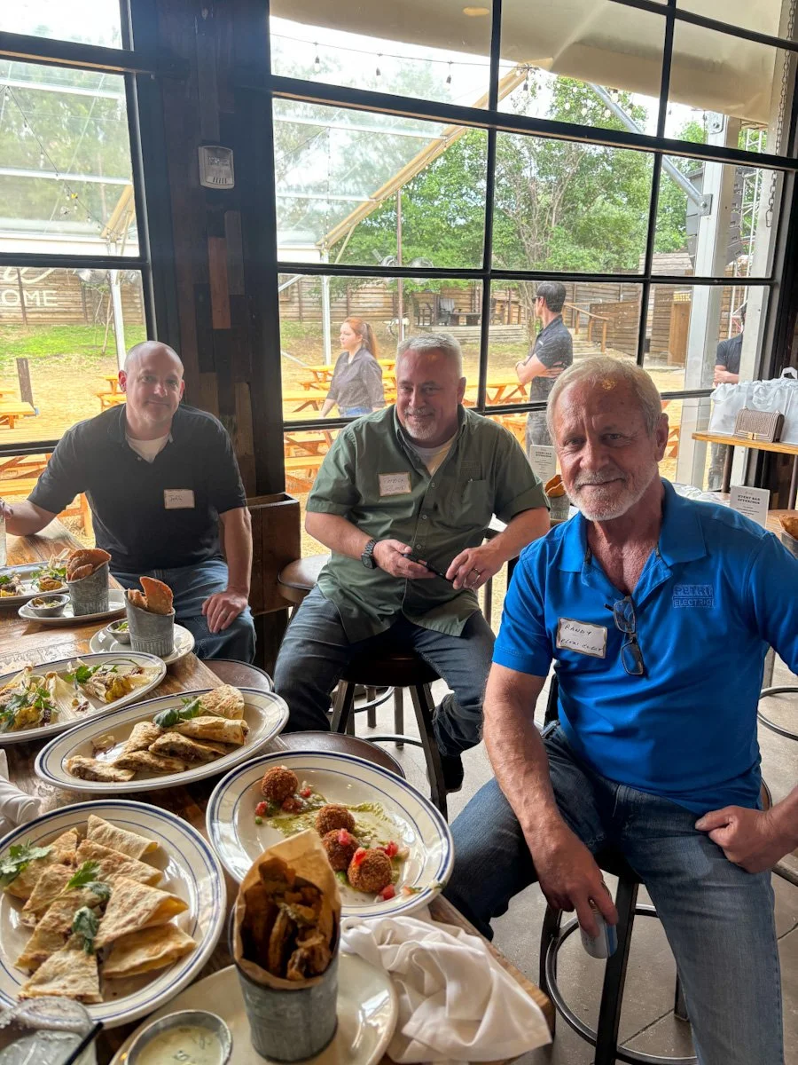Three men sitting at a table inside a restaurant with plates of food, with an outdoor patio visible through large glass windows behind them.