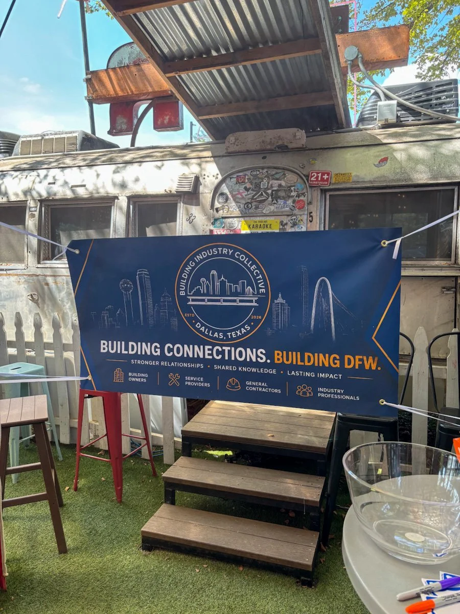 A blue banner with building industry icons and Dallas skyline illustration, hanging in front of a vintage trailer with stickers, set on grass in an outdoor area.