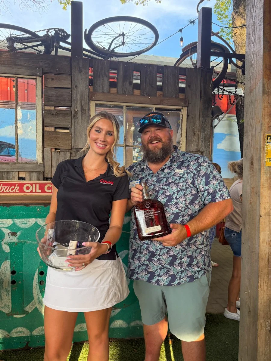 A woman and a man are smiling and posing together inside a rustic bar or restaurant. The woman is holding a clear bowl, and the man is holding a large bottle of liquor. There is a wooden wall, a window, and bicycles mounted on the roof in the background.