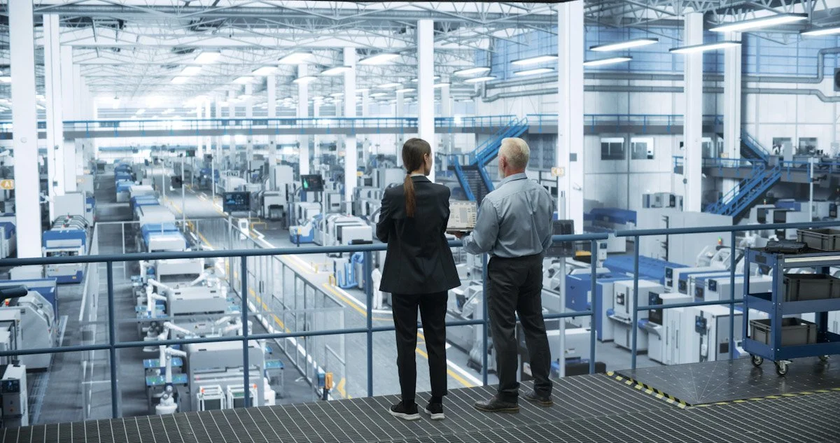 Two workers, a woman and a man, are standing on an observation platform in a large industrial factory or warehouse, looking at machinery and conveyor belts below.