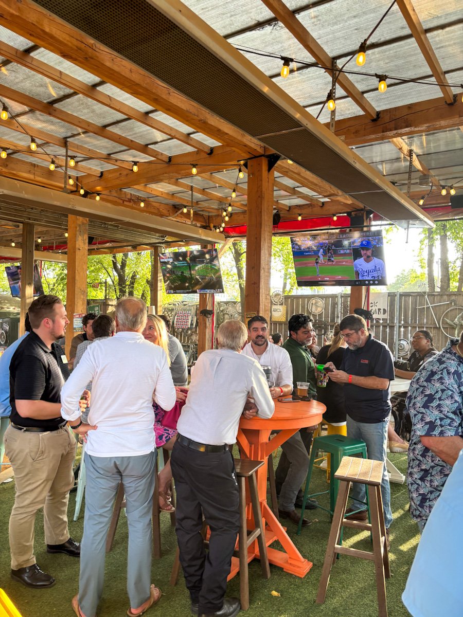 People gathered at a bar or outdoor lounge area with string lights and multiple TV screens showing a baseball game.