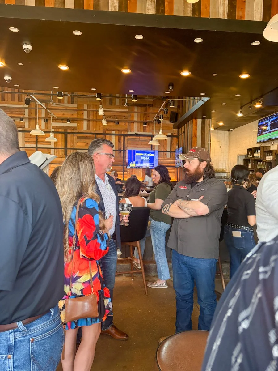 People socializing in a bar or restaurant setting with wooden decor, some holding drinks, and a TV screen in the background displaying sports.