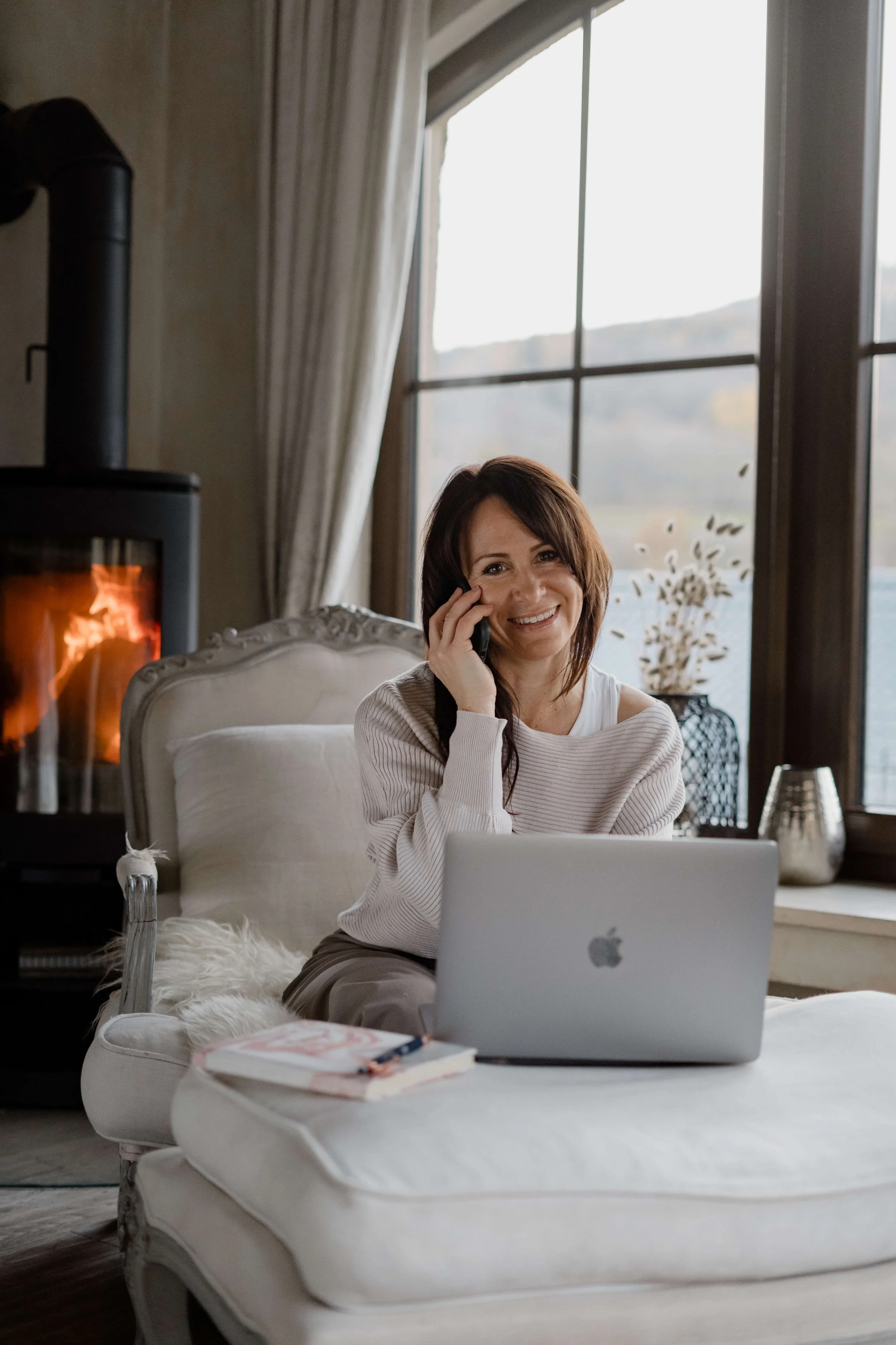 Frau sitzt auf einem weißen Sessel, spricht am Telefon, vor einem Laptop, im Hintergrund ein Kamin mit Feuer, große Fenster mit Blick auf die Berge und einen See.