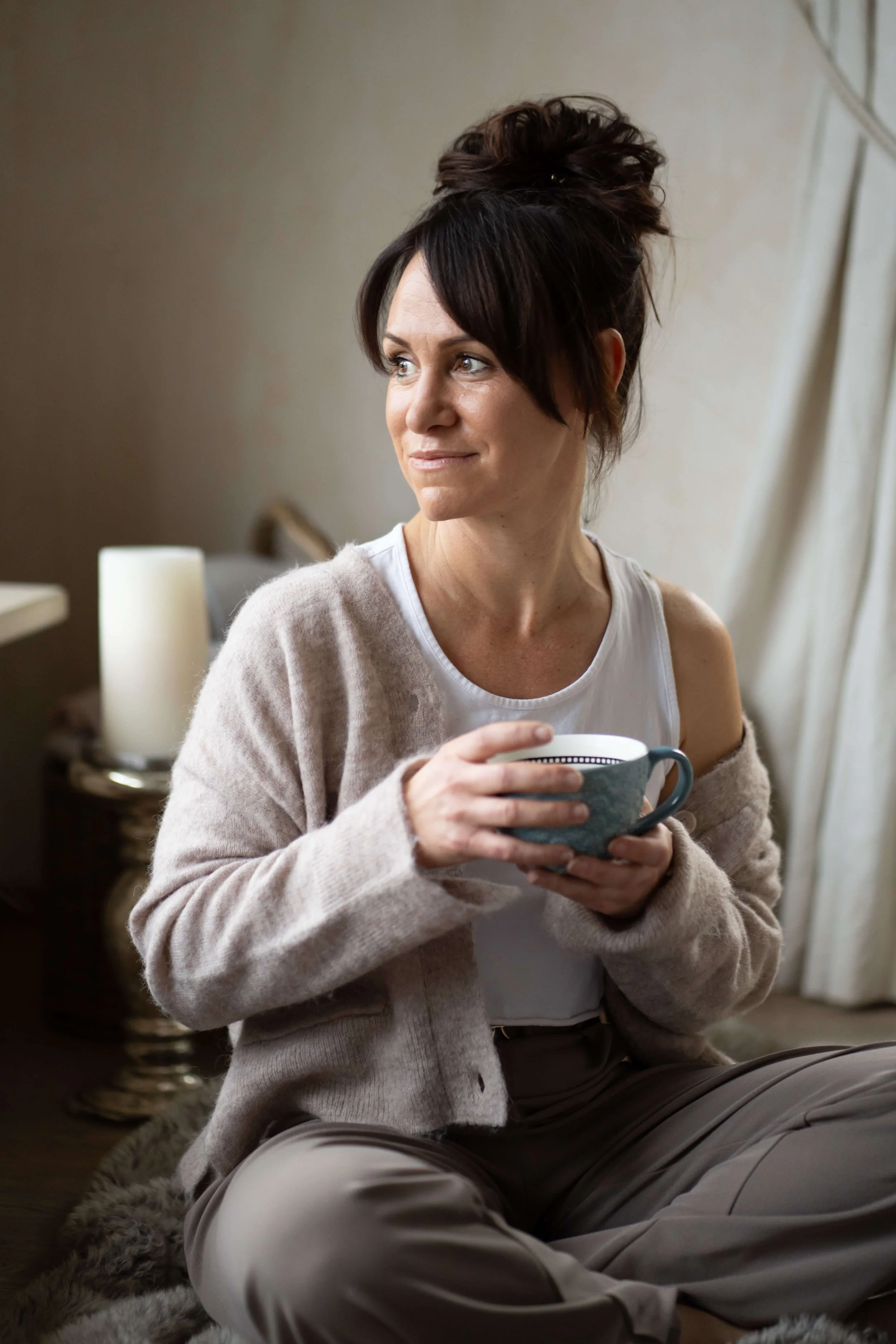Eine Frau sitzt im Raum, hält eine Tasse und sieht nach draußen, mit einem freundlichen Ausdruck.