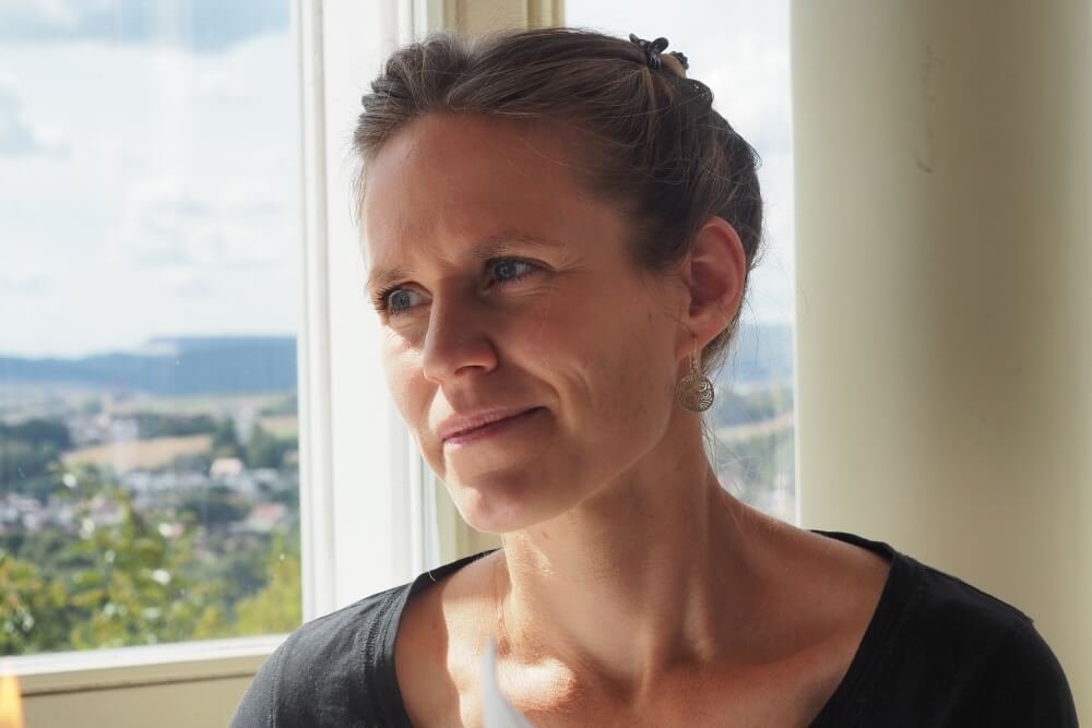 Nahaufnahme einer Frau mit dunklem Haar, die vor einem Fenster mit Blick auf die Landschaft steht.