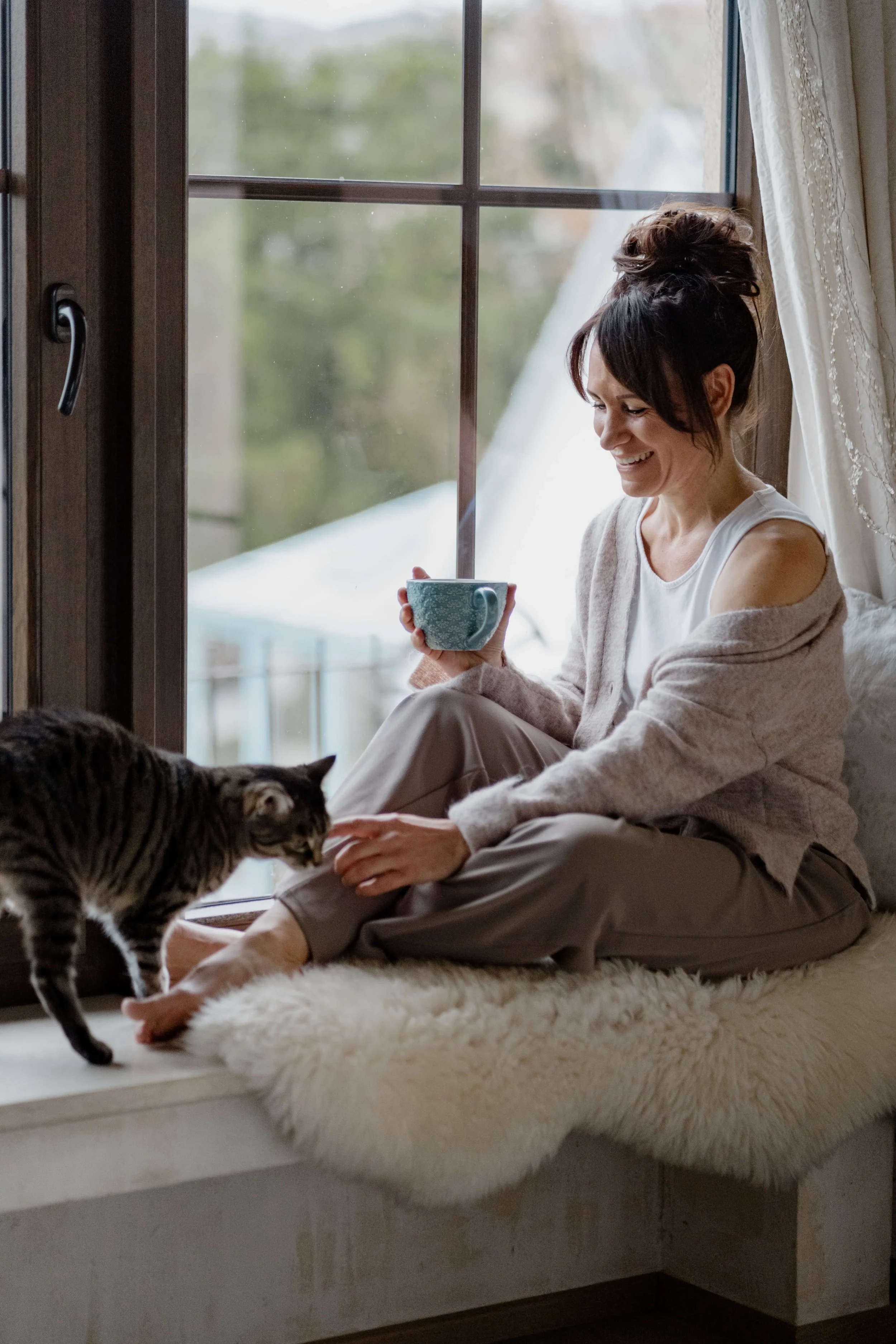Frau sitzt am Fenster mit einer Tasse, streichelt eine Katze, lächelt, bei Tageslicht