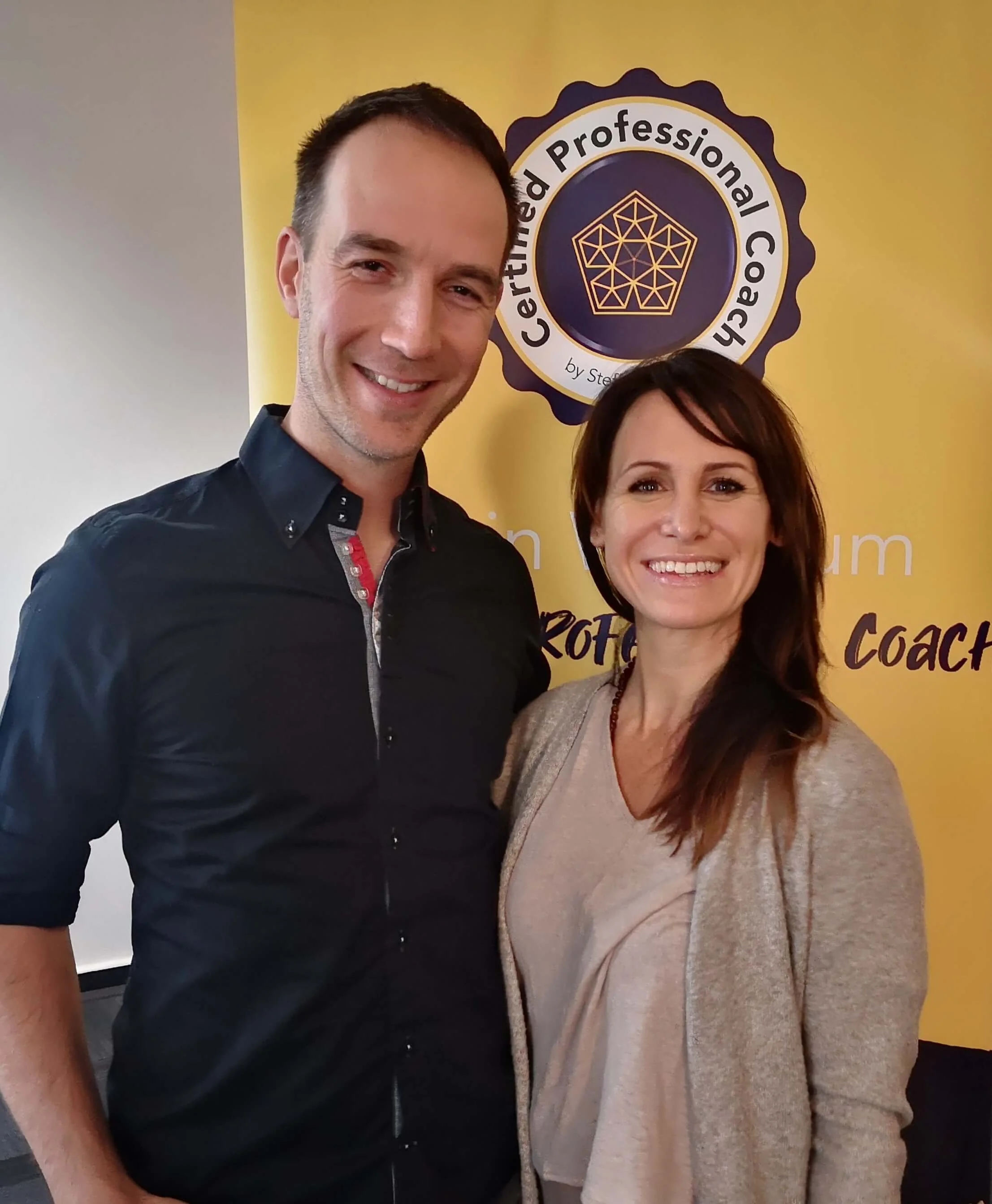 Ein Mann und eine Frau stehen vor einem gelben Hintergrund mit einem Logo, das 'Certified Professional Coach' und eine geometrische Form zeigt. Beide lächeln in die Kamera.